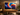A large flat-screen television mounted on a wall in a modern living room, displaying a vivid swirling abstract wallpaper in blue, orange, yellow, and purple. The TV sits above a long wooden media console, flanked by speakers, with framed wall art, plants, and a coffee table in the room.