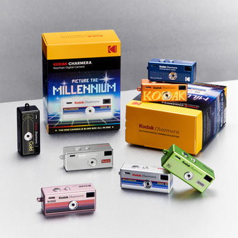 Seven Charmera Millennium keychain cameras in different colours arranged around their yellow blind boxes