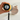 Hand holding the yellow camera by its strap, round screen facing a pink alarm clock
